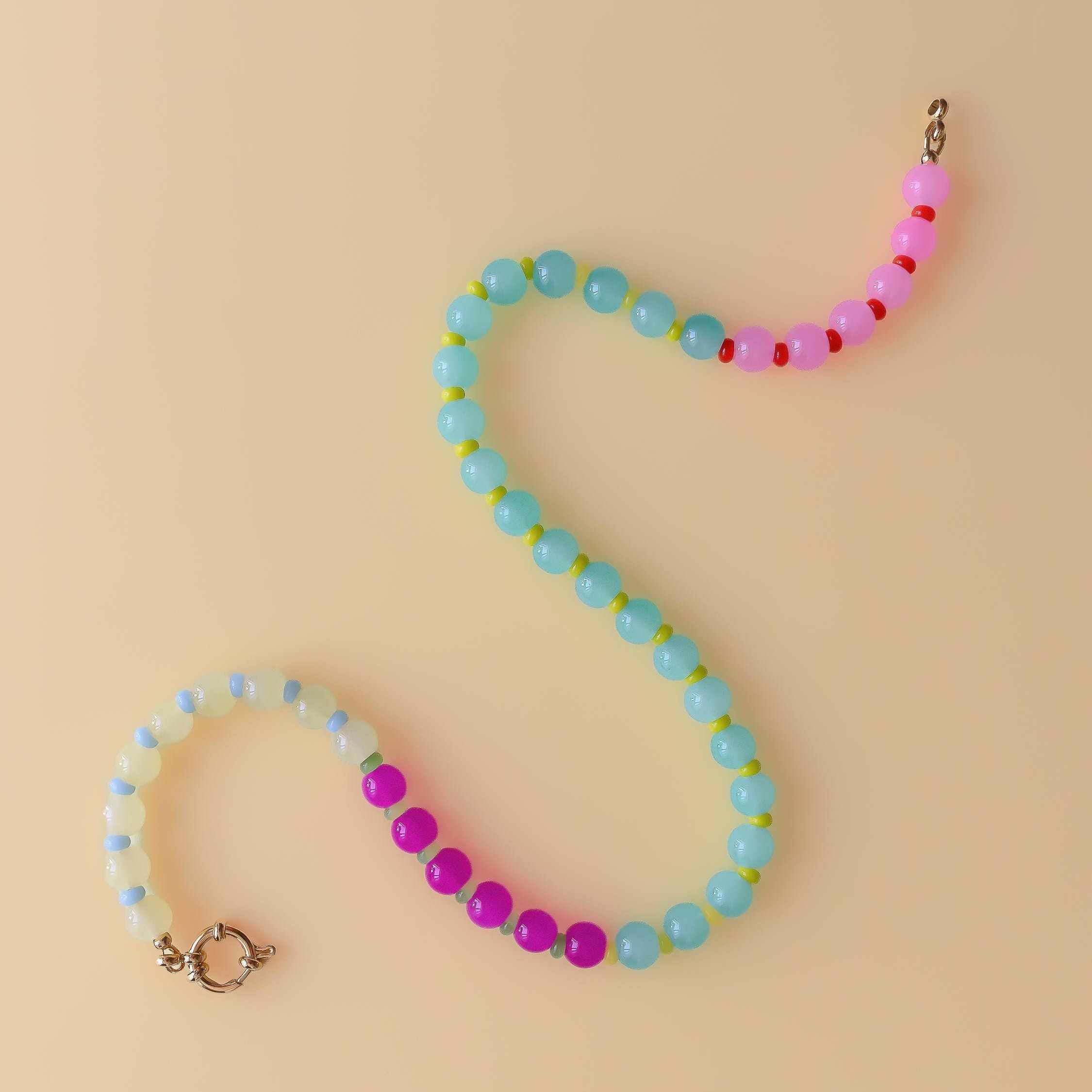 The Tutti Frutti necklace laid in a long S-curve on a pale peach background, from pale lemon beads at the gold clasp, through magenta and a long run of aqua, to candy pink at the far end